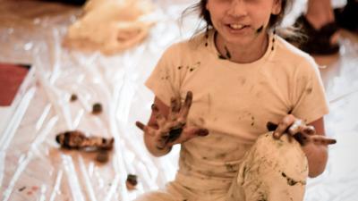 Child in paint-splattered clothes sits on floor, smiling with painted hands, surrounded by tubes in a messy, creative space